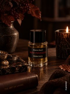 Sur une table en bois, dans une lumière chaude et tamisée, une bouteille du parfum « Musc Ravageur » est posée à côté d'une bougie allumée, de livres et de feuilles d'automne.