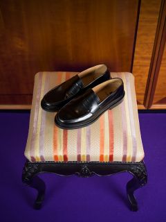 Une paire de mocassins en cuir noir repose sur un tabouret rembourré rayé avec des pieds noirs ornés, qui se trouve sur un tapis violet.