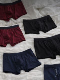 Six paires de boxers pour hommes en noir, bordeaux et bleu marine sont étalées sur un fond en tissu blanc.