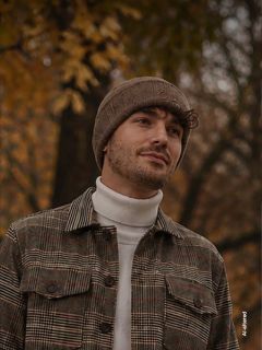 Un homme avec un bonnet marron et une veste à carreaux se tient à l'extérieur devant un fond d'arbres d'automne, regardant sur le côté et souriant légèrement.