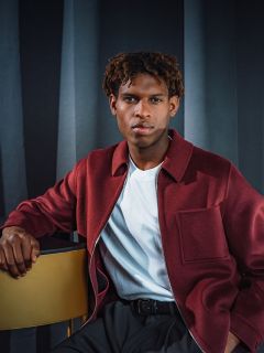 Un homme en veste marron et chemise blanche est assis à côté d'une table dorée, posant devant des rideaux sombres.