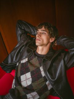 Une personne aux cheveux bouclés, vêtue d'une veste en cuir et d'un pull argyle, se détend sur un fauteuil rouge, penchée en arrière, les mains croisées derrière la tête.