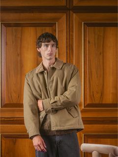 Un homme en veste beige se tient devant une porte lambrissée et regarde la caméra avec un visage impassible.