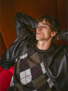 Sur un fond en bois, un homme en veste de cuir et pull argyle est penché en arrière, les mains derrière la tête, regardant vers le haut.