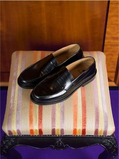 Une paire de mocassins en cuir noir repose sur un tabouret rembourré rayé, derrière lequel se trouve un mur en bois.