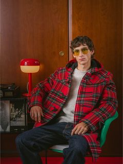 Un homme en manteau à carreaux rouges et lunettes de soleil jaunes est assis sur une chaise verte, avec une lampe rouge et un mur en bois en arrière-plan.