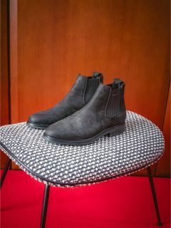 Une paire de bottines Chelsea noires sur une chaise à motifs noirs et blancs devant un tapis rouge et un mur en bois.