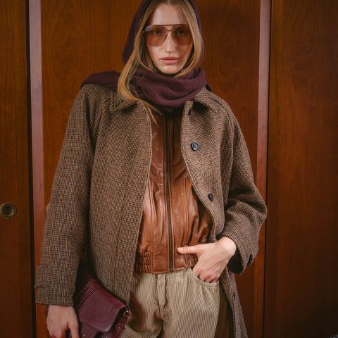 Une femme avec des lunettes de soleil, une écharpe marron, un manteau et une veste en cuir, tenant un sac à main et se tenant devant un fond en bois.