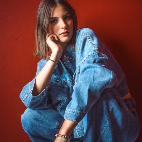 Une femme en veste et jean en jean est assise devant un mur rouge vif, le menton appuyé sur la main, regardant la caméra.