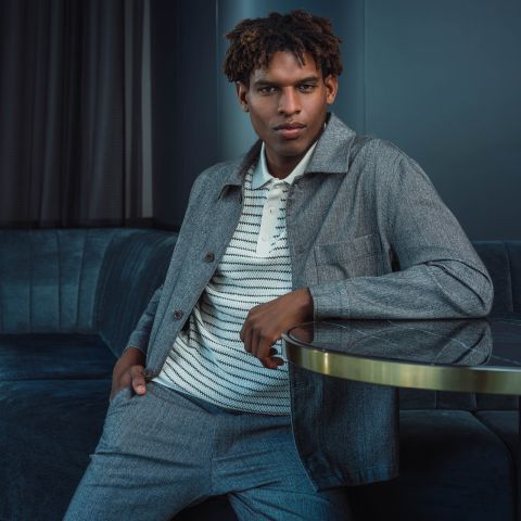 Un homme en costume gris et chemise rayée est assis sur un canapé bleu, un bras appuyé sur une table basse en verre, regardant la caméra avec assurance.