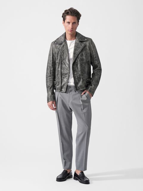 Une personne vêtue d'une veste en cuir grise, d'une chemise blanche, d'un pantalon gris et de mocassins noirs se tient devant un simple fond blanc.