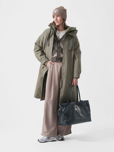 Une femme vêtue d'un manteau vert, d'un pantalon à motifs, de baskets et d'un bonnet tient un grand sac texturé et se tient devant un simple fond blanc.