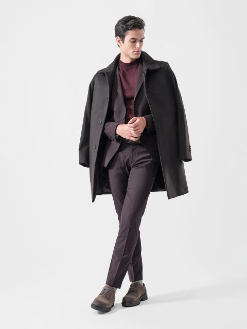 Un homme portant un manteau foncé sur les épaules, un costume bordeaux, un pull bordeaux et des chaussures marron, debout devant un fond clair et uni.