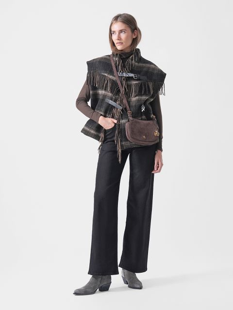 Une femme vêtue d'un poncho à carreaux, d'un haut marron, d'un pantalon noir et de bottes grises, portant un sac à bandoulière marron, se tient devant un fond blanc.