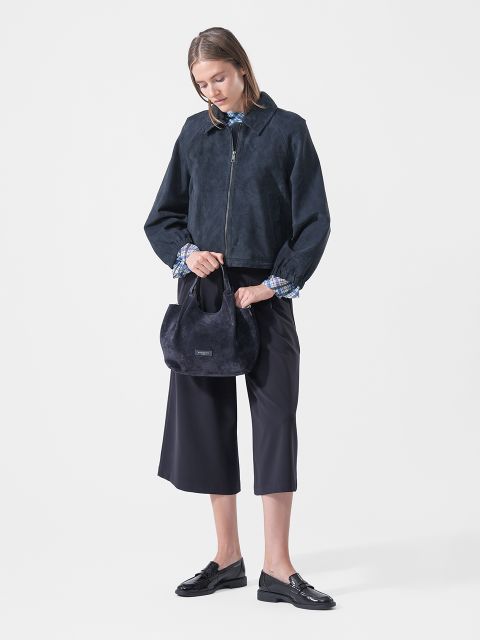 Une femme vêtue d'une veste bleu marine, d'un pantalon culotte et de mocassins, tenant un petit sac à main bleu marine, se tient devant un simple fond blanc.