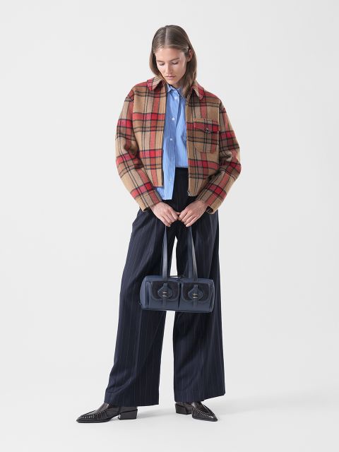 Une femme vêtue d'une veste à carreaux, d'une chemise bleue et d'un pantalon à fines rayures larges tient un sac à main bleu marine à deux compartiments et se tient devant un simple fond blanc.