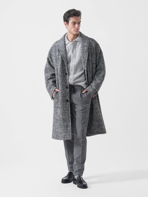 Un homme portant un manteau gris texturé, une chemise boutonnée gris clair, un pantalon gris et des chaussures noires se tient les mains dans les poches devant un fond blanc.
