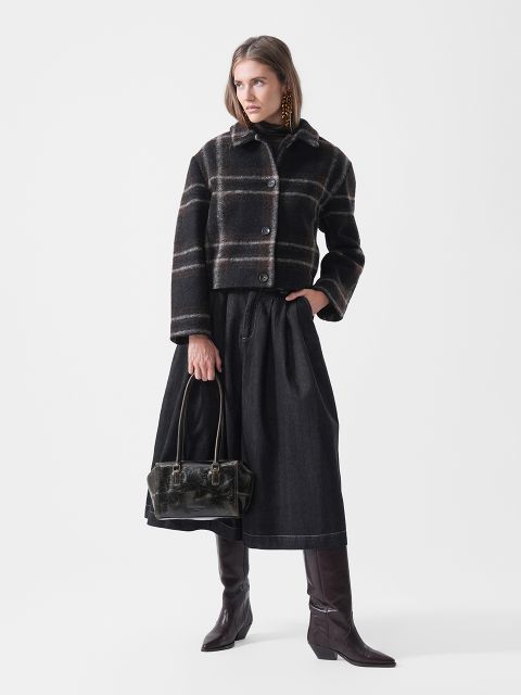 Une femme vêtue d'une veste à carreaux, d'une jupe noire et de bottes hautes marron, tenant un sac à main noir, se tient devant un simple fond blanc.