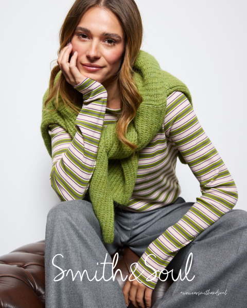 Une femme en chemise rose et cardigan avec un pantalon blanc pose devant un fond blanc. Le texte indique « Smith & Soul » en bas.