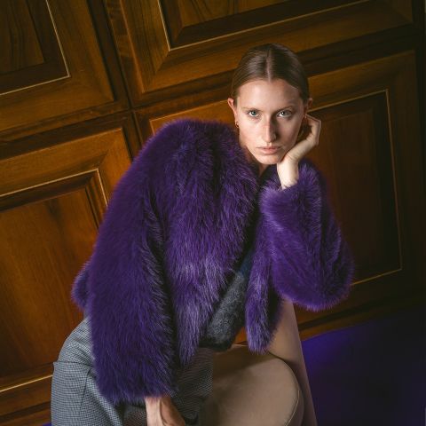 Une personne vêtue d'un manteau en fausse fourrure violette est appuyée contre une chaise, regardant l'appareil photo, avec des murs lambrissés en arrière-plan.