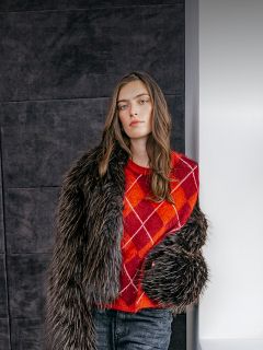 Une femme porte un pull argyle rouge-orange sous une veste en fausse fourrure foncée.