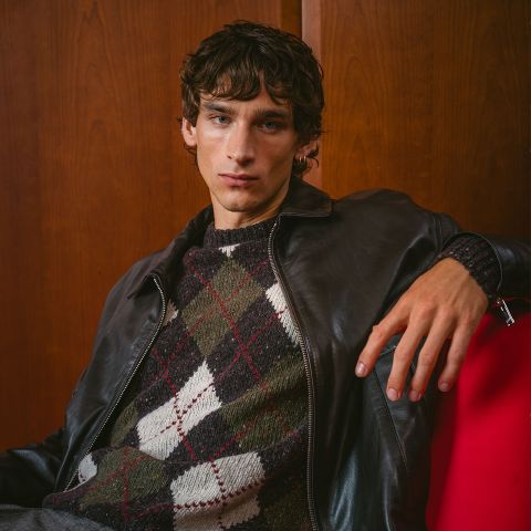 Un jeune homme vêtu d'une veste en cuir foncé et d'un pull Argyle est assis sur une chaise rouge, posant devant un mur en bois et regardant l'appareil photo.