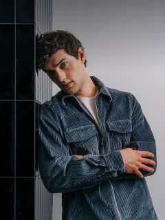 Un homme portant une chemise en velours côtelé bleue zippée, les bras croisés, appuyé contre un mur carrelé sombre.