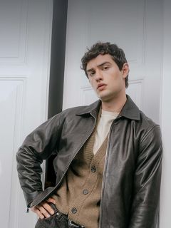 Un homme portant une veste en cuir foncé sur un cardigan beige clair et une chemise crème, appuyé contre un cadre de porte.