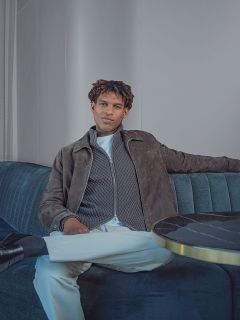 Un homme assis sur un canapé, portant une veste zippée grise, une chemise blanche et un pantalon blanc.