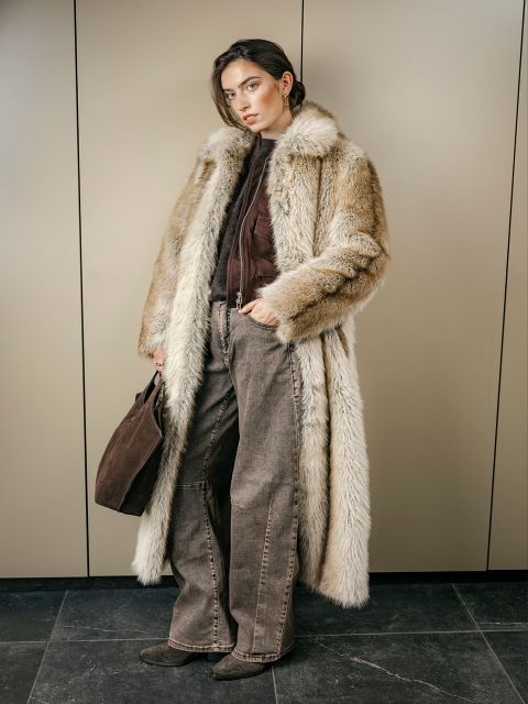 Une femme vêtue d'un long manteau de fourrure, d'un pantalon marron et de bottes se tient dans une pièce, tenant un sac à main marron et regardant la caméra avec un visage inexpressif.