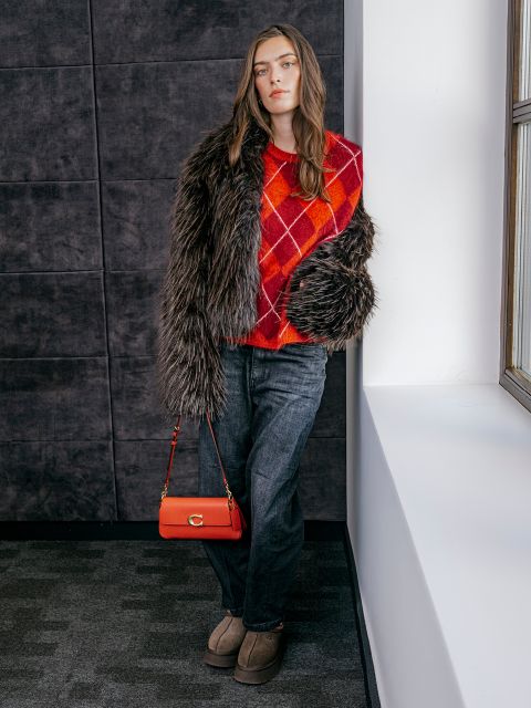 Une femme vêtue d'un pull rouge à losanges, d'un manteau en fausse fourrure, d'un jean gris et de chaussures marron clair est appuyée contre un mur près d'une fenêtre, tenant un sac à main rouge.