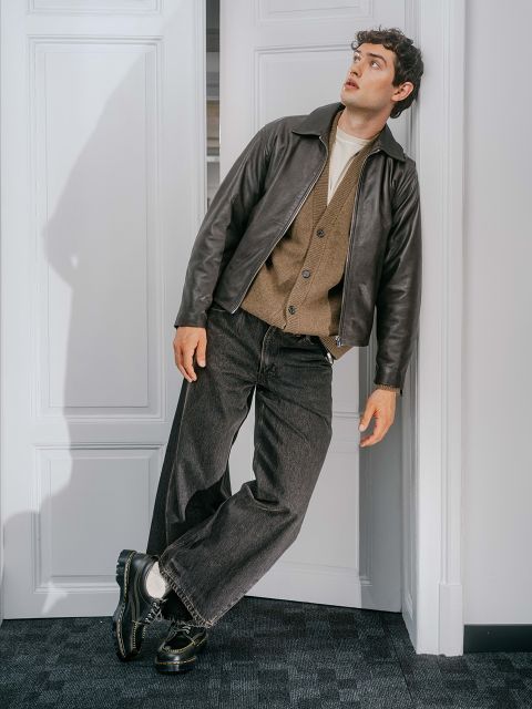 Un homme en pantalon foncé, veste marron et manteau de cuir est appuyé contre une porte blanche et regarde vers le haut avec une expression surprise.
