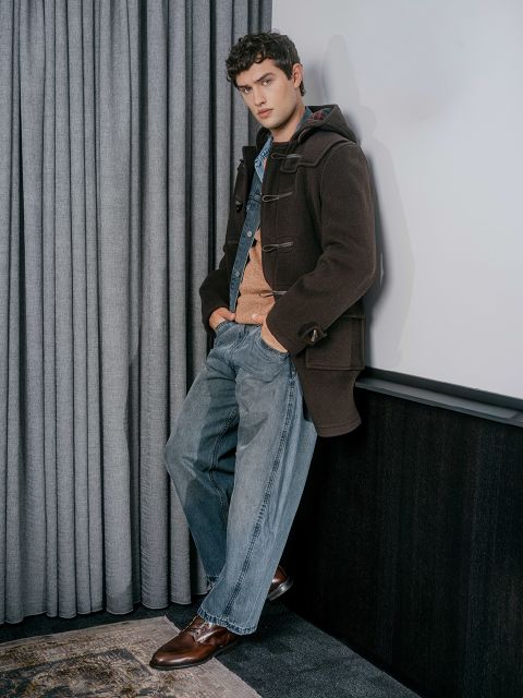 Un jeune homme en manteau marron, pull orange et jean bleu est appuyé contre un mur, les mains dans les poches, à côté de rideaux gris.