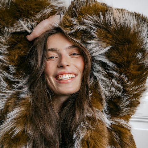 Femme souriante vêtue d'un manteau en fausse fourrure marron et blanc, les cheveux longs détachés, posant avec une main au-dessus de la tête devant un fond clair.