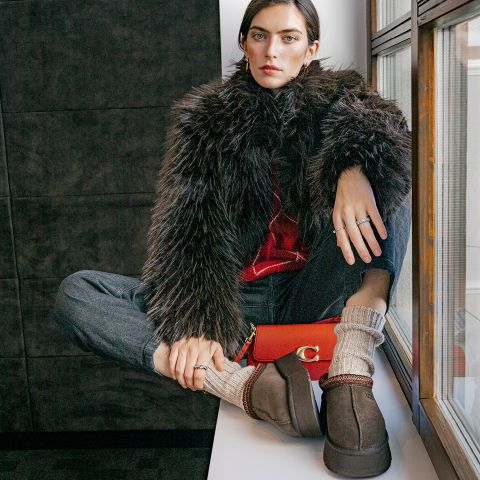 Une femme vêtue d'un manteau en fausse fourrure, d'un jean et de chaussettes douillettes est assise à la fenêtre, tenant un sac orange et regardant directement l'appareil photo.