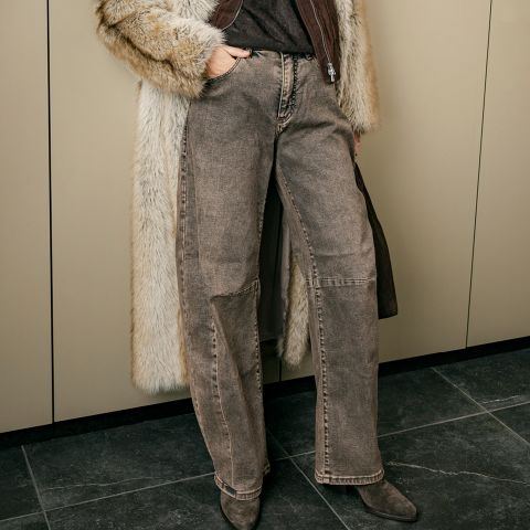 Une personne portant un jean marron à jambe large, un manteau de fourrure et des bottes pointues, tenant un sac, se tient devant des armoires beiges.