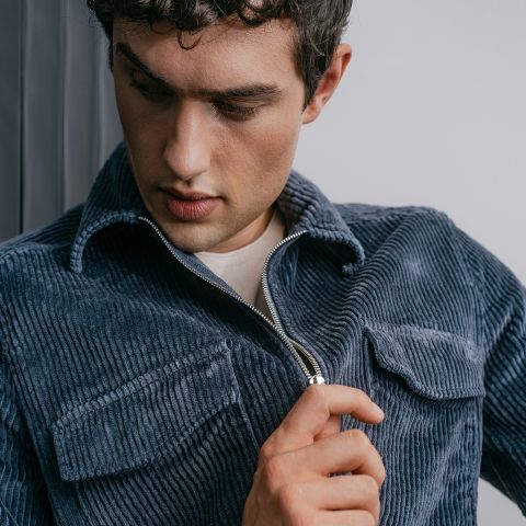 Un homme aux cheveux courts et bouclés, vêtu d'une veste en velours côtelé bleue sur une chemise blanche, regarde vers le bas tout en fermant la fermeture éclair de sa veste.