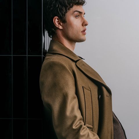 Un homme en manteau marron se tient de profil devant un mur noir et blanc, regardant vers l'avant avec une expression sérieuse.