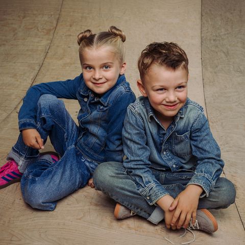 Deux enfants en tenue de jean sont assis sur un plancher en bois et sourient à la caméra. L'un porte des baskets roses, l'autre des chaussures marron clair.