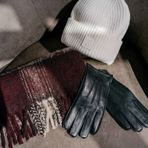 Un bonnet côtelé blanc avec un logo carré, une écharpe à carreaux marron châtaigne avec des franges et une paire de gants en cuir noir.