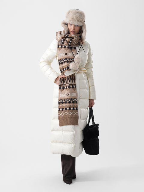 Une femme porte un long manteau blanc en duvet, une écharpe à motifs bruns avec le logo UGG et une chapka en fausse fourrure ; elle tient un sac à main noir à la main.