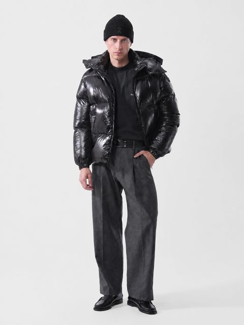 Un homme portant un bonnet noir, une doudoune noire brillante, une chemise noire, un pantalon gris et des mocassins noirs.