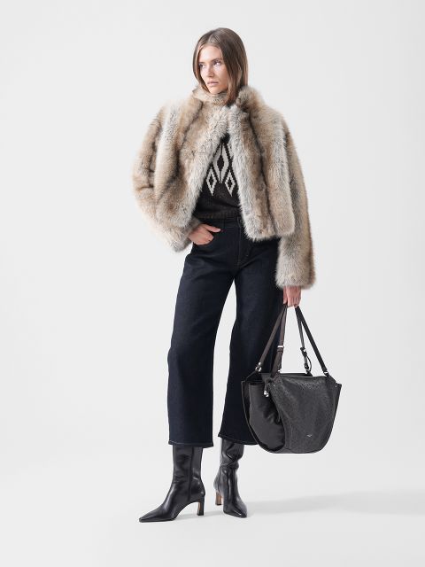 Un mannequin porte une veste en fourrure sur un pull à motifs, un jean foncé à jambes larges et des bottes noires à talons, et porte un grand sac à bandoulière noir.