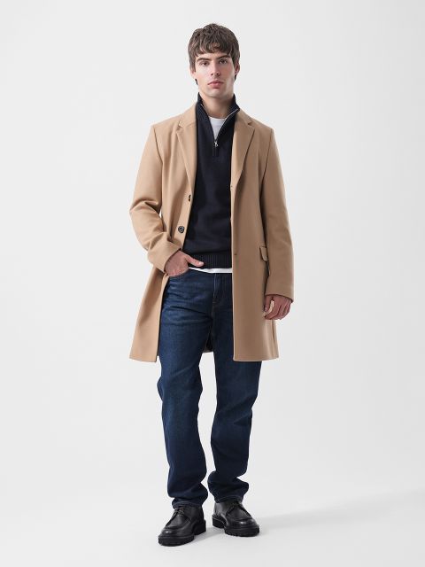 Un homme portant un manteau beige, un pull bleu marine à col zippé, un t-shirt blanc, un jean bleu foncé et des chaussures noires.