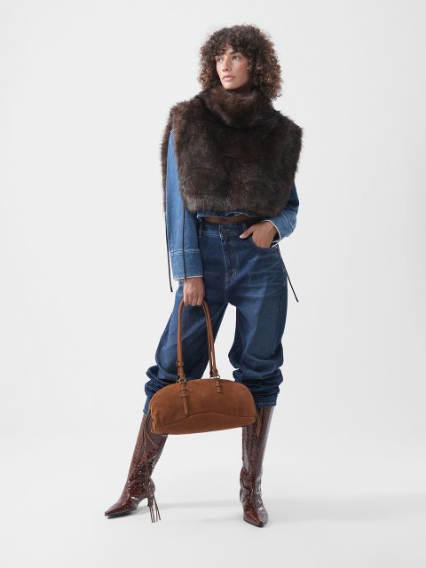 Une personne portant une veste en jean, un jean foncé et une écharpe en fourrure foncée, tenant un sac à main en daim marron et portant des bottes hautes en cuir gaufré marron.