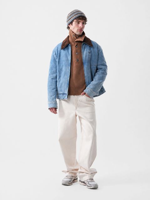 Un homme portant une veste en jean bleu sur un pull boutonné marron, un pantalon crème et une casquette.