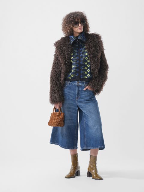 Une personne portant une veste en fausse fourrure marron, une veste à motifs zippée, un jean culotte bleu et des bottines vert olive, tenant un petit sac à main tressé marron.