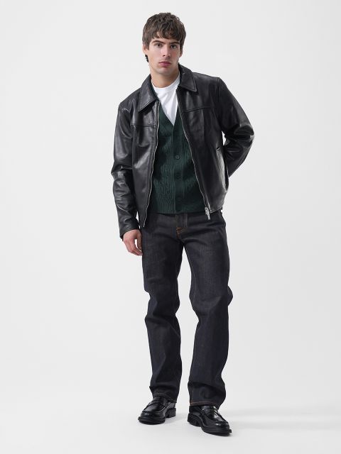 Un homme portant une veste en cuir noire sur un cardigan vert, un t-shirt blanc, un jean foncé et des mocassins noirs.