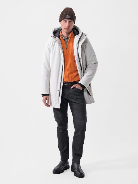 Un homme portant une parka blanche à capuche, un pull orange à col zippé, un jean noir et un bonnet marron.