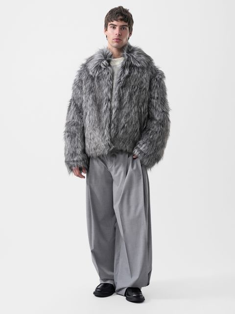 Un homme portant une veste en fausse fourrure grise, un pull crème et un pantalon gris à jambes larges.